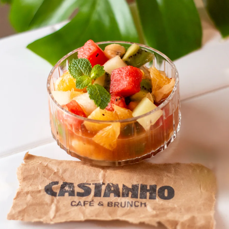 Pote de vidro canelado com salada de frutas do Castanho — melancia, kiwi, laranja, uva e maçã, com folhas de hortelã — sobre o guardanapo de papel com a marca da casa